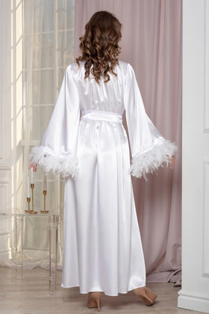 Woman wearing a white satin robe with feathered cuffs in a softly lit room.