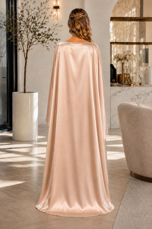 A woman stands with her back to the camera wearing a flowing, floor-length draped garment in a warm beige or champagne tone, photographed in a sunlit indoor room with large windows and a fireplace.