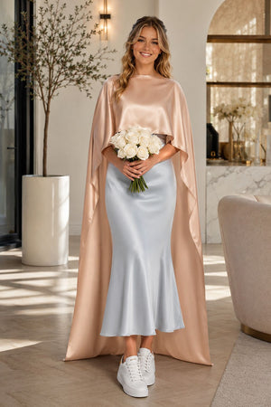 A woman models a floor-length satin gown with a draped cape-style overlay in warm beige and dark brown, styled in a sunlit indoor room with a fireplace and window.