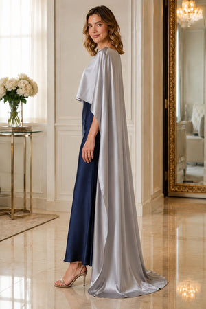 A woman models a floor-length evening gown in a luxurious indoor setting. The dress features a navy blue satin skirt and a flowing silver cape overlay with wide draped sleeves.