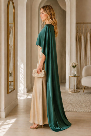 A woman stands in profile in a softly lit boutique interior, wearing a floor-length champagne satin gown with a dramatic draped emerald green cape. She holds a small textured clutch and wears nude heels.