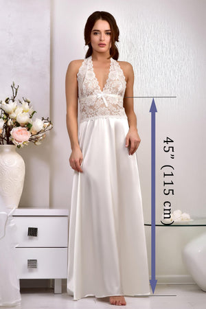 A woman modeling a sleeveless, backless A-line wedding dress with a lace bodice and satin skirt, standing in a brightly lit room with a white side table and flowers in the background.