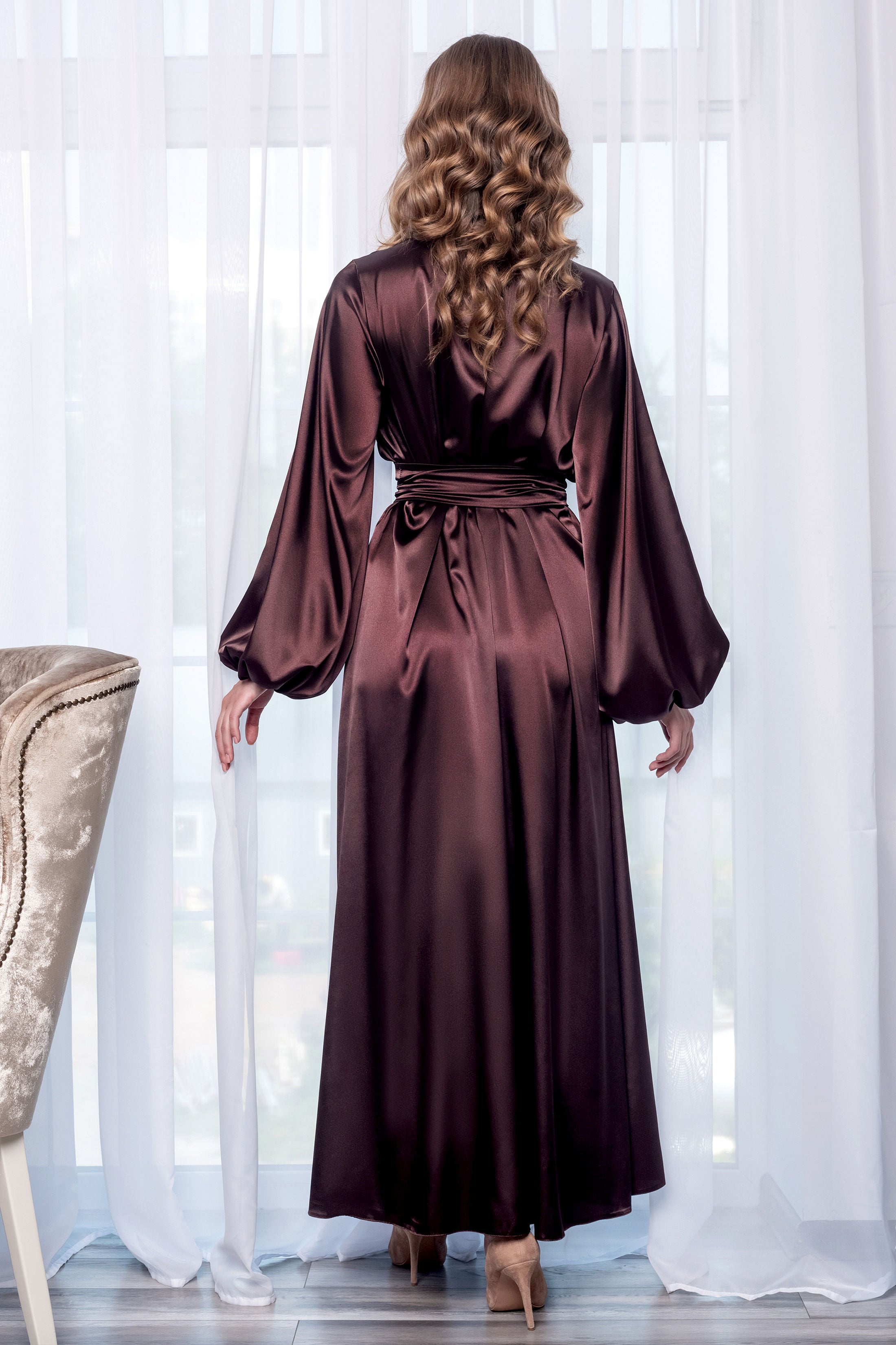 A woman viewed from behind wearing a long, floor-length satin robe in a deep burgundy-brown color, standing in front of white sheer curtains.