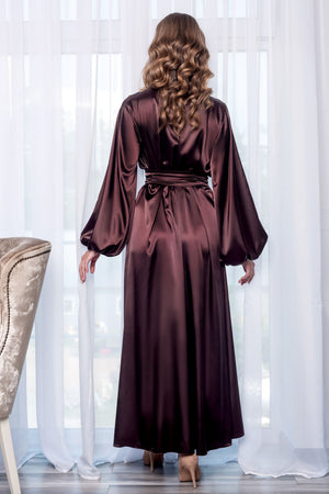 A woman viewed from behind wearing a long, floor-length satin robe in a deep burgundy-brown color, standing in front of white sheer curtains.