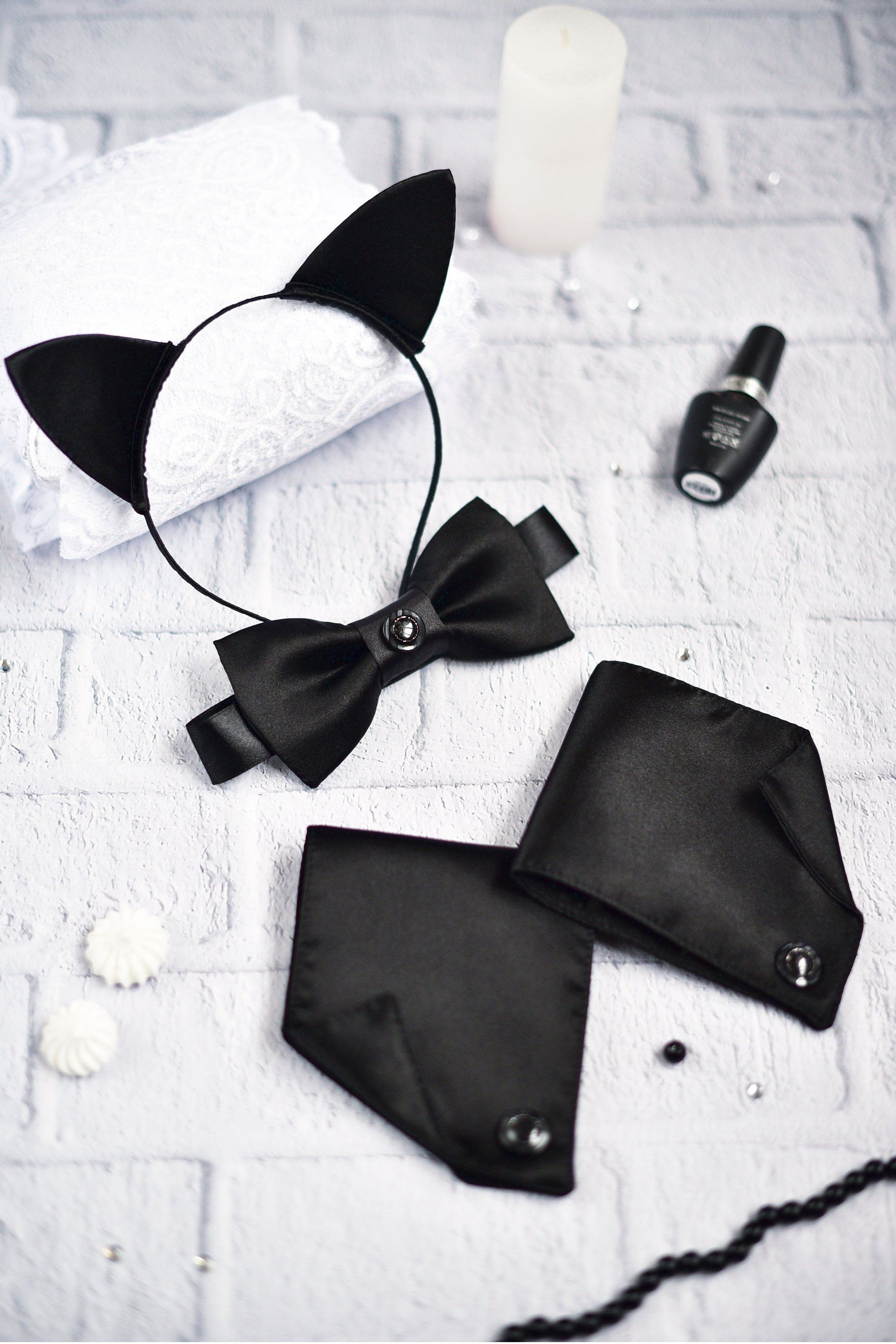 A black satin cat ears cosplay set displayed on a white surface, including a pair of cat ears, two cuffs, and a bow tie, with a small bottle and a candle nearby.