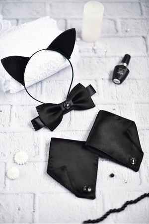A black satin cat ears cosplay set displayed on a white surface, including a pair of cat ears, two cuffs, and a bow tie, with a small bottle and a candle nearby.