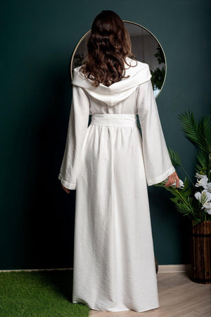 Woman wearing a long white coat standing in front of a mirror against a dark green wall.