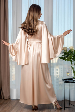 Woman in a long, light pink satin robe standing in a room with large windows.