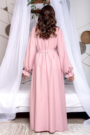 Woman wearing a pink dress standing in a room with white curtains and a bed.