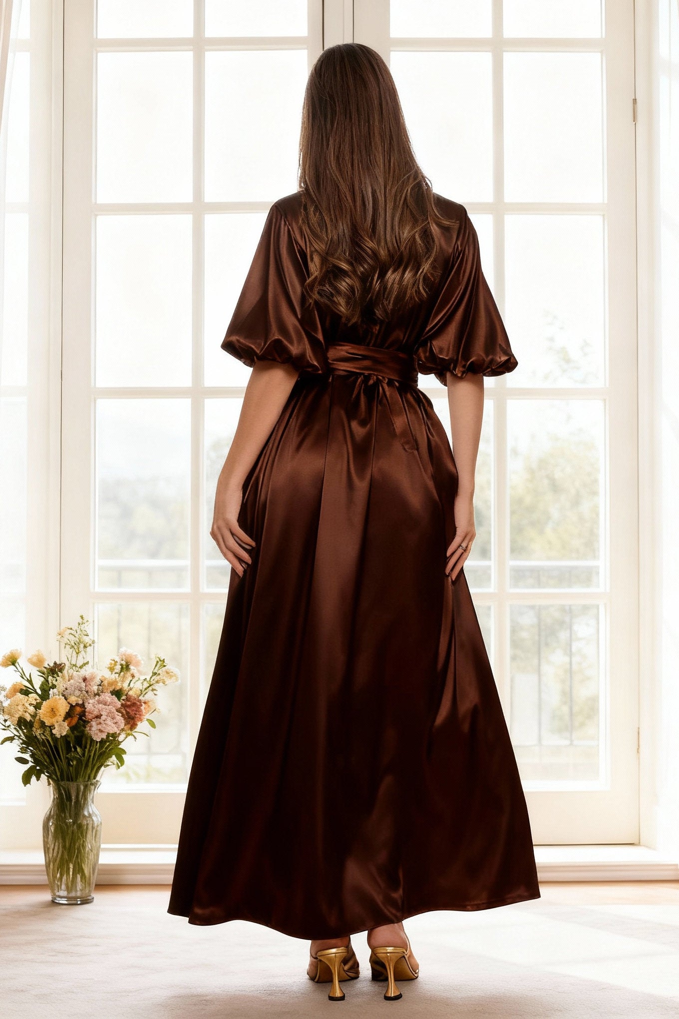 Woman wearing a brown satin dress standing in front of a large window with a vase of flowers on the floor