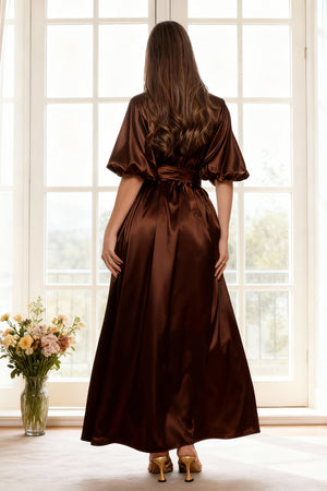 Woman wearing a brown satin dress standing in front of a large window with a vase of flowers on the floor