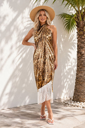 Woman wearing a gold sequin dress with fringe and a hat, standing against a white wall with a palm tree.
