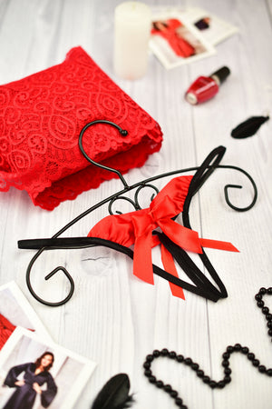 A black open string panty with a red bow, displayed on a wooden hanger, surrounded by red lace, nail polish, and other lingerie items.