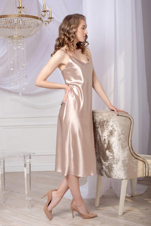 Woman in a beige satin dress standing in a room with a chandelier and chair.