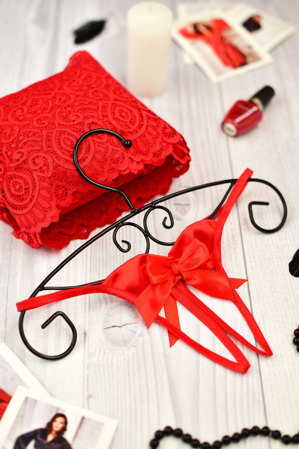 A sheer black open crotch string with satin bows, displayed on a white wooden surface with red lace, nail polish, and other accessories.