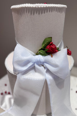 A white wedding cake topped with a red chiffon ribbon bow.