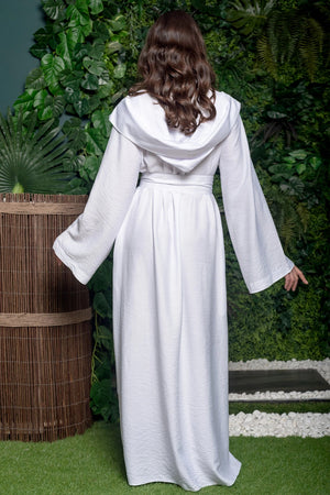 Person wearing a white hooded garment standing in a garden setting with greenery and a wooden barrel.