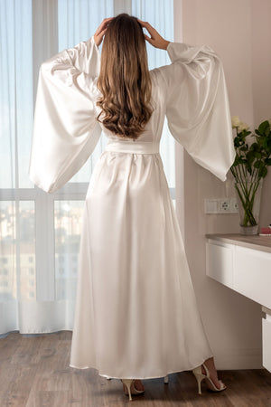 Woman in a long white dress standing in a room with a window and a vase of flowers