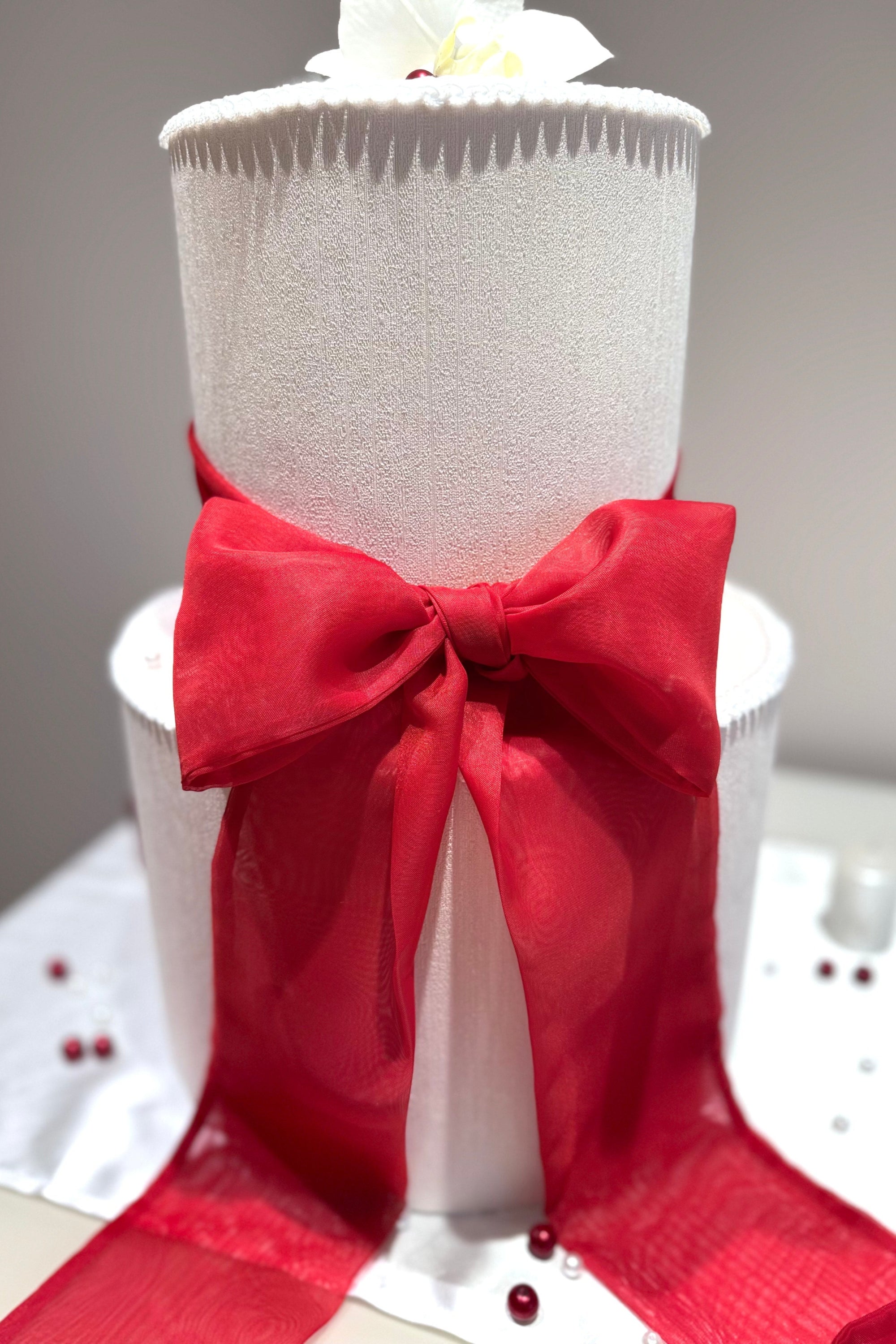 A chiffon ribbon bow in red, attached to a white wedding cake, set against a neutral background.