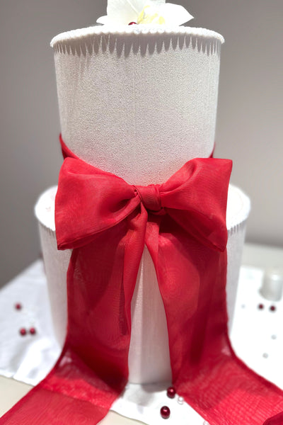 A chiffon ribbon bow in red, attached to a white wedding cake, set against a neutral background.