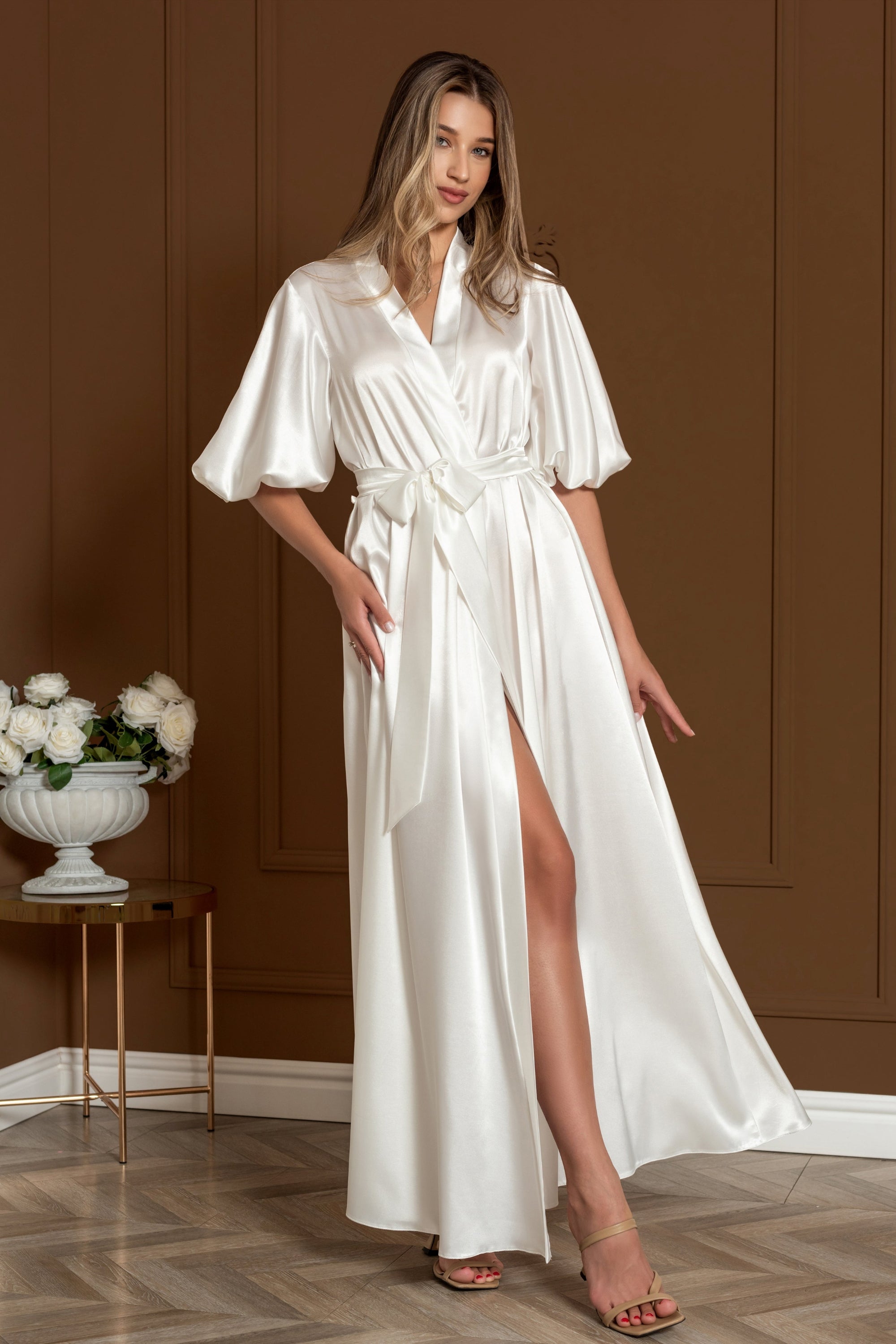 Woman wearing a white satin robe standing in a room with a brown wall and wooden floor.