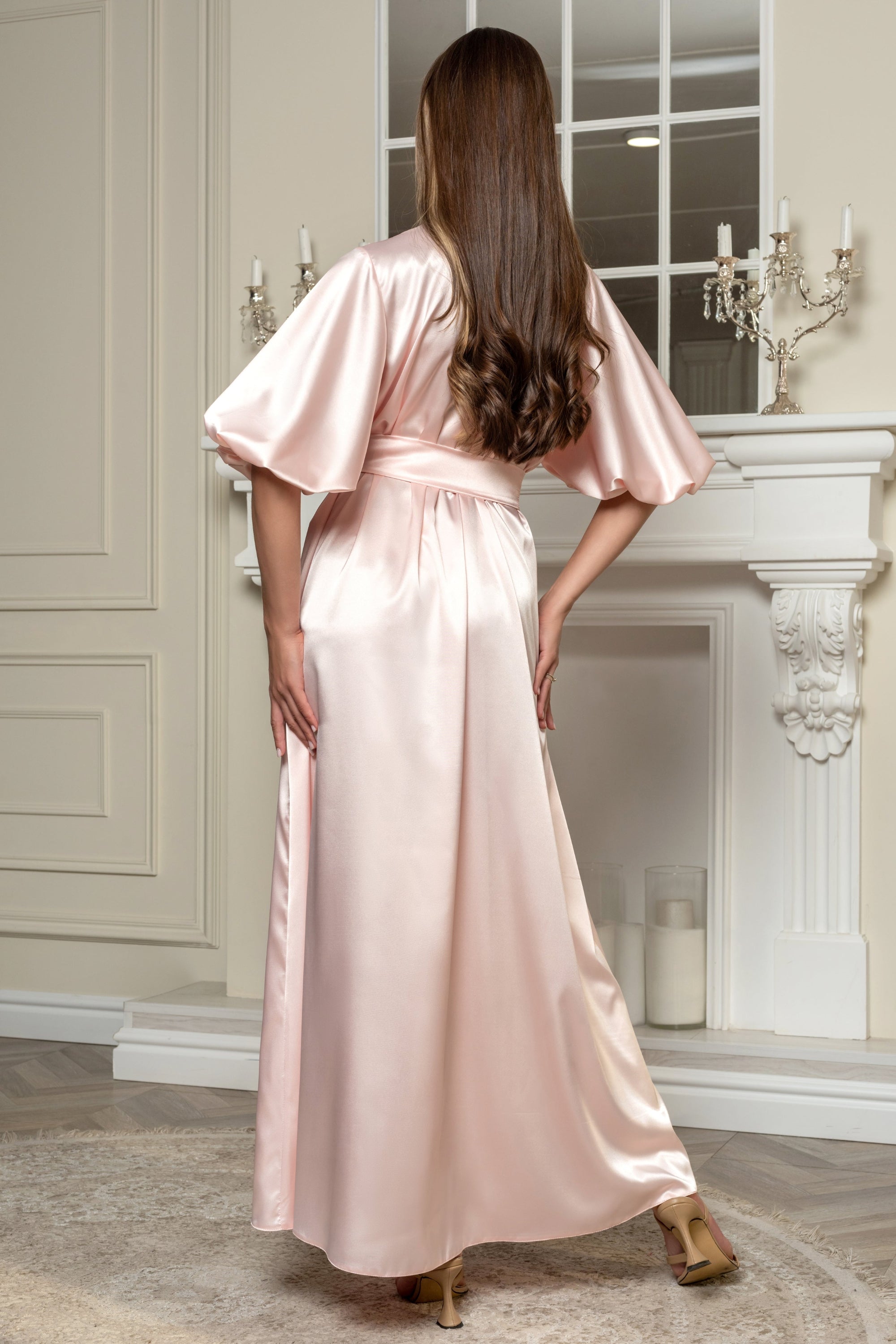 Woman in a pink satin robe standing in a room with a fireplace.
