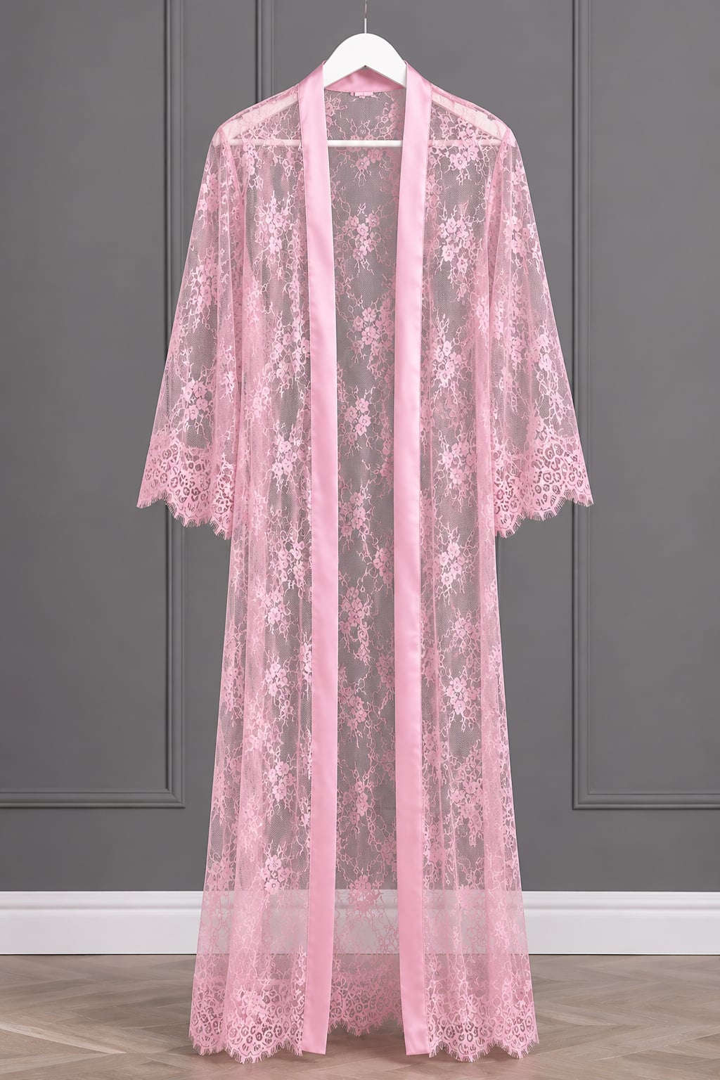 A pink sheer lace robe with floral embroidery and a matching pink satin front panel, hanging on a white hanger against a gray paneled wall.