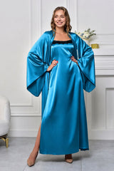 A woman modeling a floor-length satin robe in a bright teal-blue color, styled with a matching wide-sleeved cape over a black slip dress, standing in a white room with a fireplace.