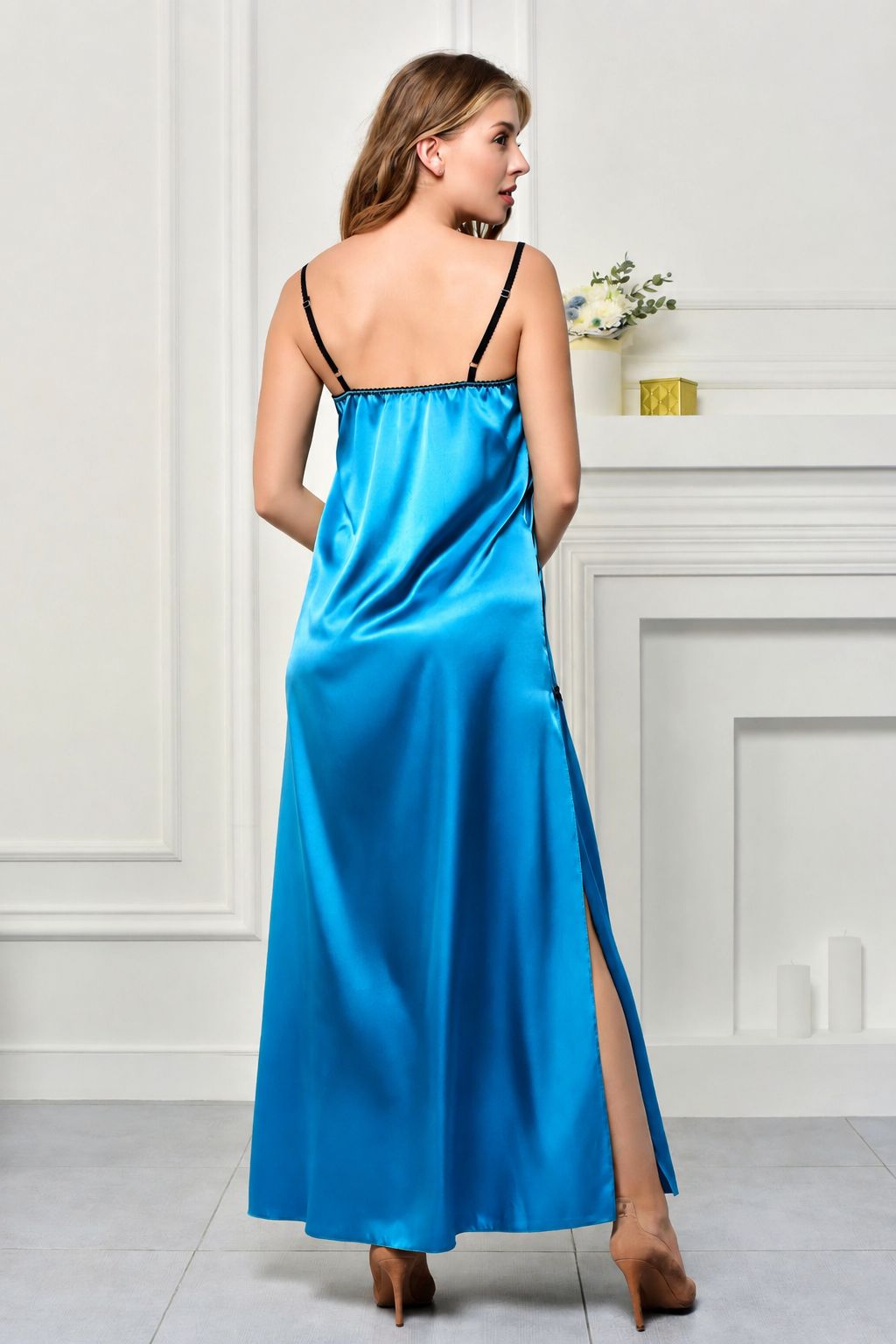 A woman wearing a bright blue satin slip dress with black spaghetti straps, shown from the back in a bright, white-paneled room.