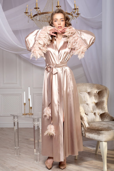 A woman in a beige satin robe with feather trim on the sleeves and belt, standing in a room with a luxurious decor.