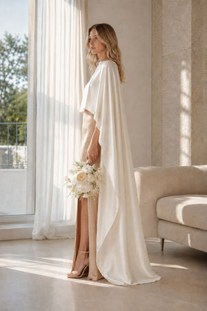 A woman stands in a bright, sunlit room wearing a flowing ivory bridal gown with a draped cape-style overlay and a high slit, holding a bouquet of white and beige flowers.