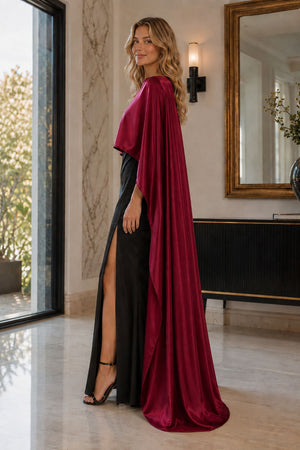 A woman models a floor-length evening gown with a high slit, black satin-like skirt, and a dramatic draped capelet in deep burgundy. She is standing in a luxurious indoor setting with marble floors, a large mirror, and a window.