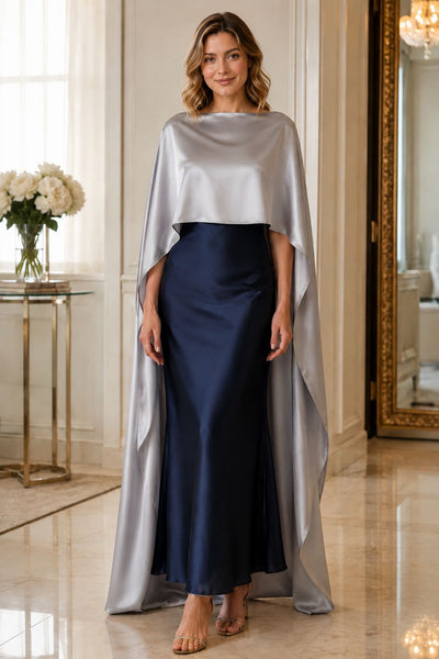 A woman models a floor-length satin gown with a silver cape-like overlay and a navy blue skirt, standing in an elegant indoor hallway with a mirror and floral table in the background.