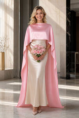 A woman models a floor-length satin gown with a high-low cape effect. The dress features a champagne beige skirt and a contrasting light pink cape overlay, set against a modern, sunlit minimalist interior.