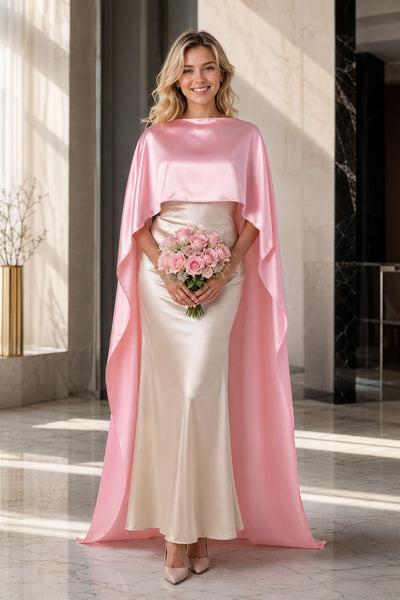 A woman models a floor-length satin gown with a high-low cape effect. The dress features a champagne beige skirt and a contrasting light pink cape overlay, set against a modern, sunlit minimalist interior.