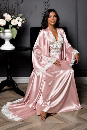 Woman in a pink satin robe with lace details sitting in a room with dark walls and a vase of flowers.