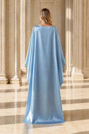 A woman viewed from behind wearing a flowing light blue satin caftan or robe with wide draped sleeves, standing in a grand hall with beige stone columns and floor.