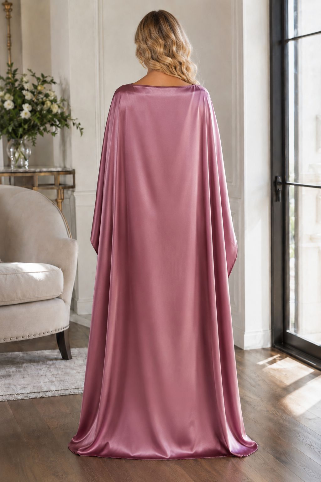 A woman stands with her back to the camera wearing a floor-length draped satin cape in dusty rose pink, set in a bright, elegant room with wood floors and a window.