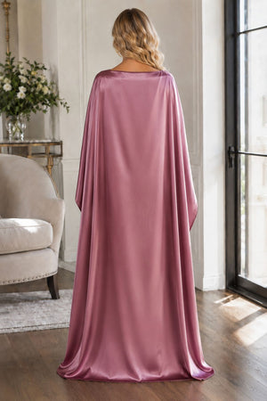 A woman stands with her back to the camera wearing a floor-length draped satin cape in dusty rose pink, set in a bright, elegant room with wood floors and a window.