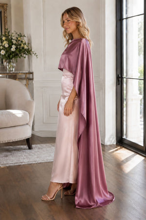 A woman models a two-piece formal gown in a bright, elegant room. The dress features a light pink satin skirt and a draped mauve-purple cape-style top with elbow-length sleeves.