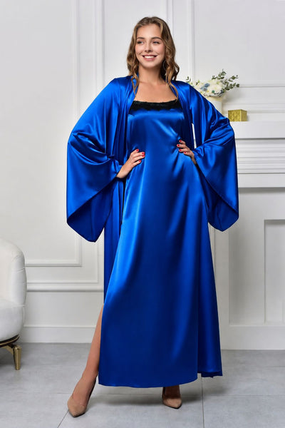 A woman modeling a royal blue satin robe with wide draped sleeves over a black slip dress, standing in a white room with a fireplace and light gray floor.