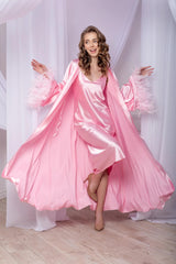 Woman in a pink satin nightgown standing in a softly lit room with white curtains.