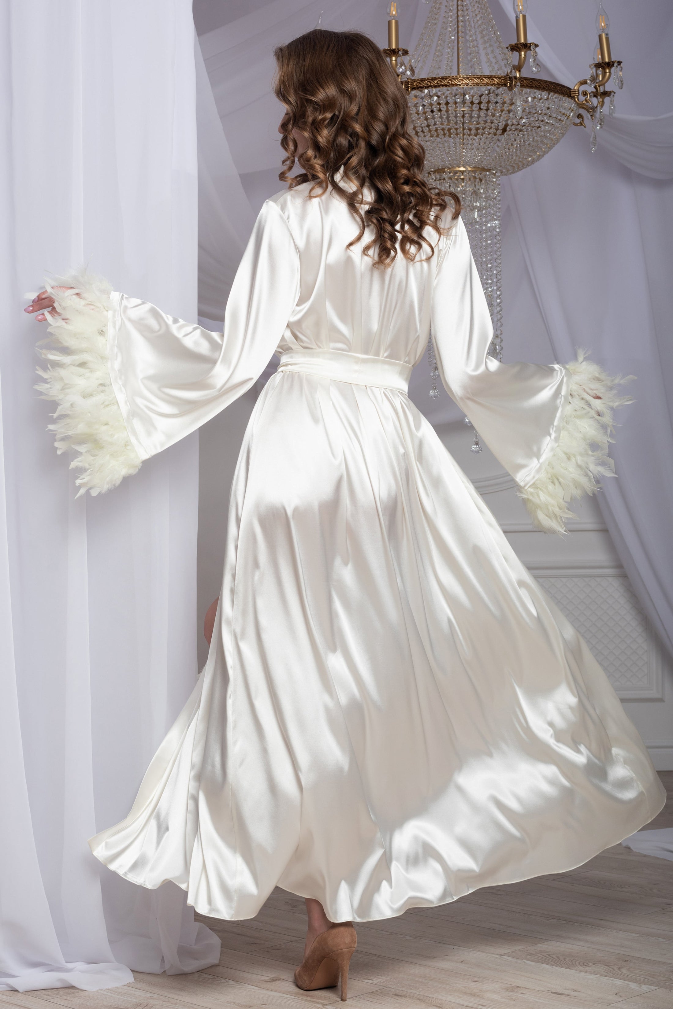 Woman in a white satin robe standing in a room with a chandelier.