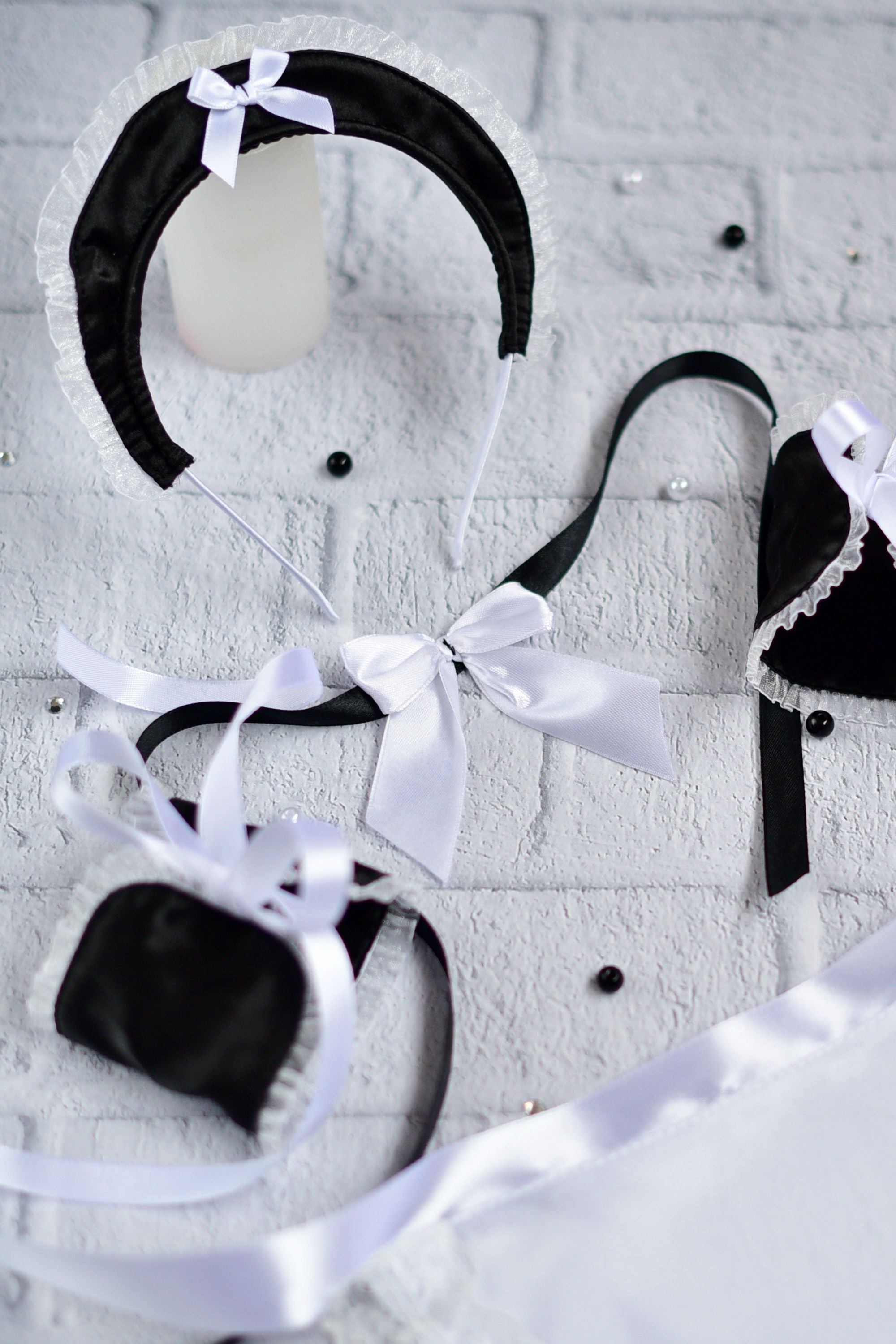 Black and white Victorian-style lingerie set including a bonnet with a white bow, a bralette, a small pouch, and matching ribbons, arranged on a white brick background.