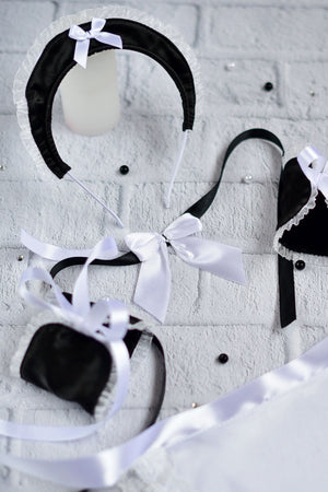 Black and white Victorian-style lingerie set including a bonnet with a white bow, a bralette, a small pouch, and matching ribbons, arranged on a white brick background.