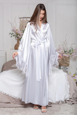 Woman wearing a white satin robe with fringe details in a bedroom setting.