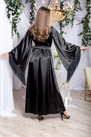 Woman in a black satin dress with fringed sleeves standing in a decorated room with white drapes and greenery.