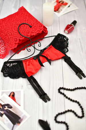 Red and black lace garter belt with attached black leg straps, displayed on a hanger over a white wood surface with red fabric, a candle, nail polish, and printed photos around it.