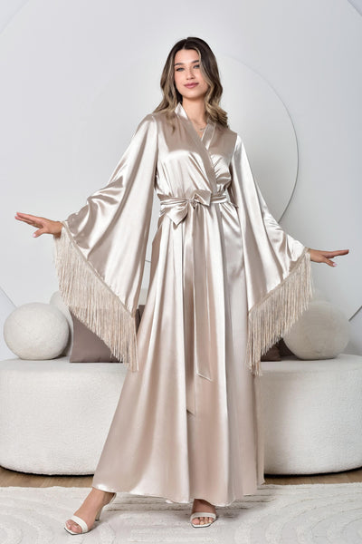 Woman wearing a long, beige satin dress with fringe details in a minimalistic setting.