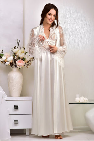 Woman in a long white lace dress standing in a room with a vase of flowers and a table.